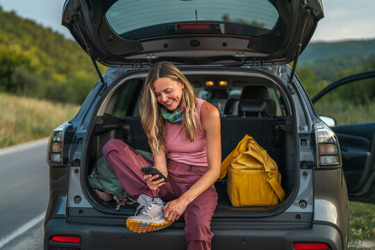 Woman smiling sitting in car trunk using smartphone