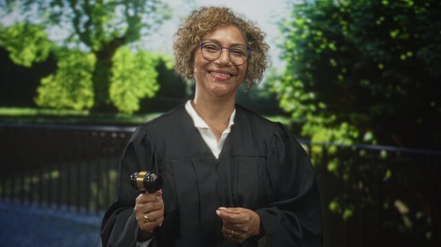 Woman judge holding gavel and smiling in sunlit park with trees and iron railing; authority confidence.