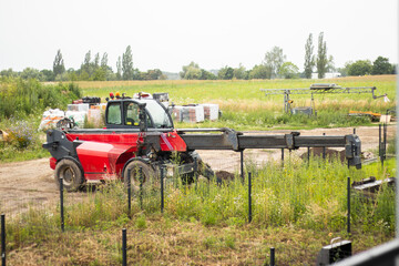 Red agricultural telehandler vehicle operating in a rural field with green grass, trees, and stacks of hay bales visible in the background © Anastasiia