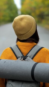 Young woman wearing a beanie and carrying a backpack with a yoga mat is walking away on a road through an autumn forest, turning her head to enjoy the peaceful scenery