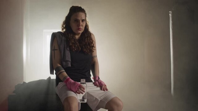 Portrait of young Caucasian female boxer sitting in smoky gym, resting after intensive training and holding water bottle. Clip is perfect for fitness marketing and motivational campaigns