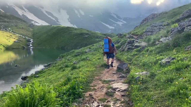 A lone tourist follows the narrow path beside Green Abudelauri Lake in Georgia, carrying camping gear through an alpine basin that represents independent travel, remote tourism, route discovery
