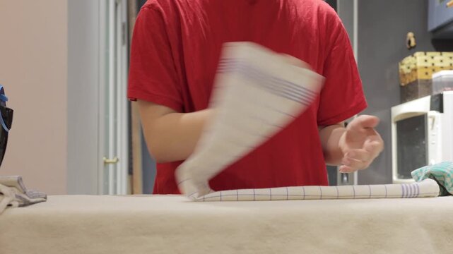 Hands fold and smooth a checked kitchen towel on an ironing board after ironing.