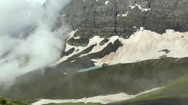 White Abudelauri Lake emerges beneath drifting fog and low clouds in Georgia's Caucasus, framed by snowfields and dark cliffs, evoking remote wilderness, alpine weather, seasonal thaw