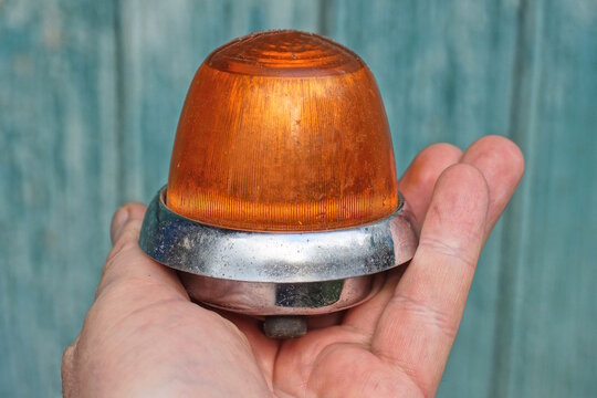 A hand holds one old rare red gray turn signal headlight from a motorcycle made of plastic and metal on a green background