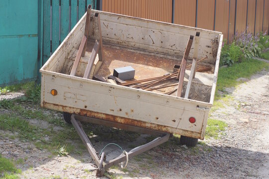 one gray open square trailer on wheels stands on the ground and grass on a summer rural day street near a fence