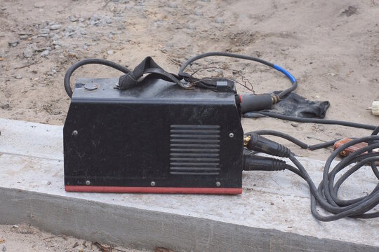 One black metal electric welding machine with wires stands on a white concrete foundation and gray ground at a construction site on a daytime street