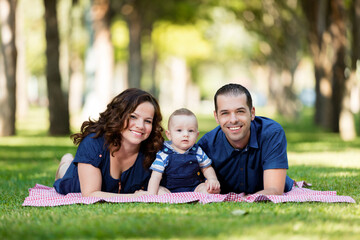 Fototapeta premium Portrait of a smiling family with a baby sitting on a blanket on the green grass in a park on a sunny day.