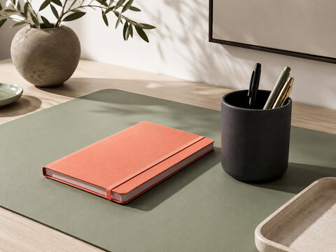 A minimalist desk setup featuring stationery items including an orange notebook, a black pen holder with pens, and a small bowl.