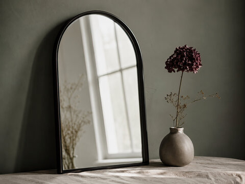 A minimalist still life featuring a black-framed mirror reflecting a room with sunlight and a small vase of dried purple flower, creating a serene and elegant composition.