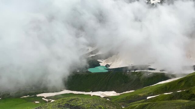 Dense mountain cloud engulfs White Abudelauri Lake under snowy Chaukhi ridges in Georgia, briefly exposing milky glacial water while illustrating rapid weather shifts, limited visibility
