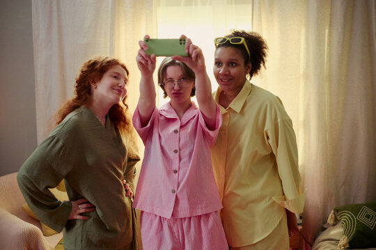 Young girls taking smartphone selfie during sleepover at home, posing together and creating social media content. Ideal for friendship, youth lifestyle, digital communication campaigns