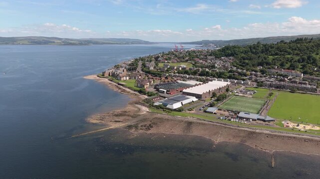 A cinematic wide aerial orbit showcasing Battery Park, the local beach, and the Firth of Clyde in Greenock, Scotland during a clear sunny day.