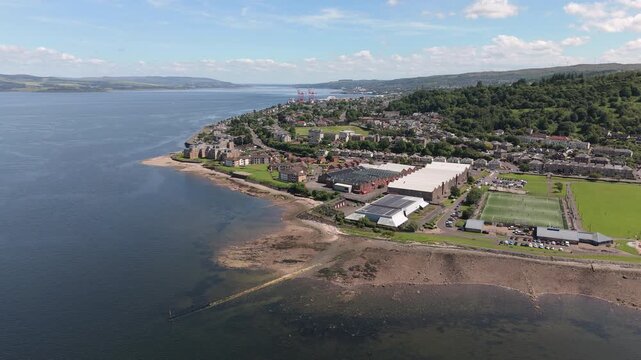 A cinematic wide aerial shot showcasing the scenic coastline of Greenock, Scotland, featuring Battery Park, local sports fields, and the Firth of Clyde under a bright blue sky.