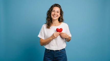 Obraz premium Beautiful Smiling Young Woman in White T-Shirt and Blue Jeans Holding Red Heart, Casual Summer Style Portrait Against Blue Background, Love and Happiness Concept, Female Model Showing Affection