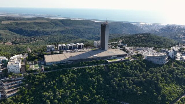 Drone flight capturing an elevated panoramic view of the iconic institutional architecture and modern skyscraper campus on Mount Carmel surrounded by green forests overlooking the Mediterranean coast