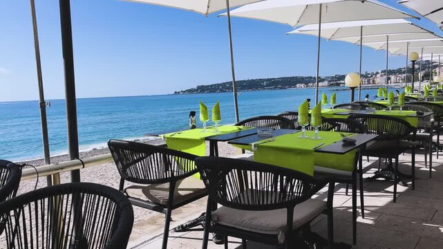 Bright outdoor restaurant terrace overlooking Plage de Fossan and the Mediterranean Sea in Menton, France. Green table settings, clear glassware, black chairs, and white umbrellas frame a scenic coast