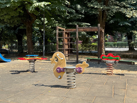 Empty playground featuring colorful spring riders. A wooden climbing frame. And a slide on rubber flooring. Surrounded by green trees and an iron fence in the background