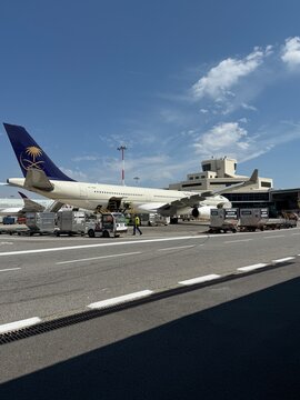 Airplane preparing for departure on the tarmac, with a ground crew loading cargo onto the aircraft beside the control tower under a sunny sky in Milan Malpensa Airport Italy 11 July 2026
