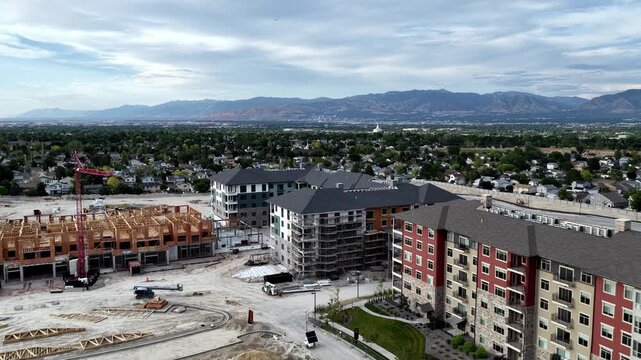 Apartment construction project beside completed multifamily buildings in Taylorsville, Salt Lake County, Utah, USA, beneath Wasatch Range with active earthworks and tower crane, drone forward flyover