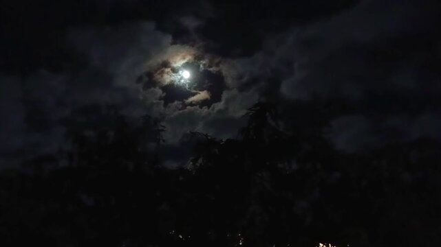 Bright full moon glows through layered clouds above dark tree silhouettes at night, with textured sky surrounding the illuminated opening and no visible people or buildings. below.