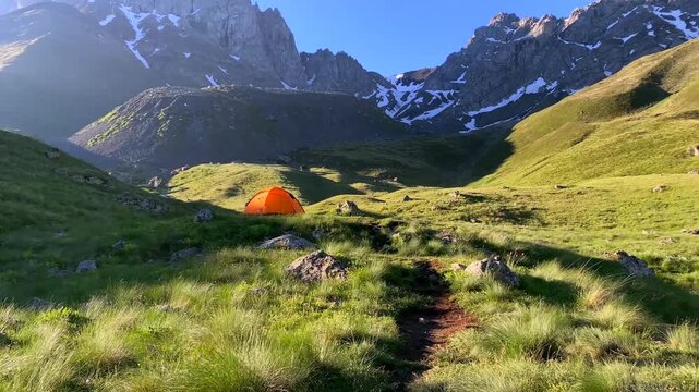 An orange tent stands in a green alpine meadow beneath the rugged Chaukhi peaks in Georgia, presenting remote camping, trekking accommodation, outdoor equipment, self-supported travel