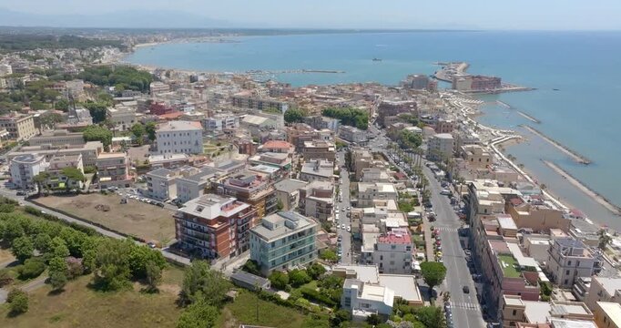 Aerial panoramic view of the sunny promenade and coastline in Anzio, in province of Rome, Italy. There are beachfront buildings, sandy shores, breakwaters, and clear sea under a bright summer sky.