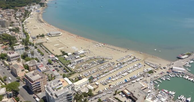 Aerial view of the coast of Anzio, in Lazio, Italy, featuring a wide sandy beach, a boat dry dock storage area, a circular harbor filled with yachts, and beachfront buildings along the clear blue sea.