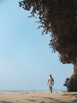 Young man walking on a sunny tropical beach under clear blue sky, rocky cliff.