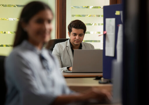 A YOUNG EXECUTIVE WORKING ON LAPTOP WITH COLLEAGUE SITTING AHEAD