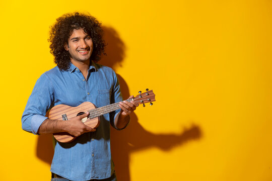 A CURLY HAIRED YOUNG MAN STANDING AND HAPPILY PLAYING UKELELE