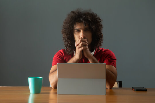 A CURLY HAIRED YOUNG MAN LOOKING AT CAMERA WHILE SITTING IN FRONT OF DIGITAL TABLET