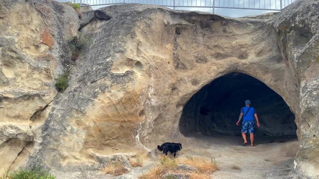 A male traveler enters a rock-cut chamber at Uplistsikhe while a dog explores nearby, showing cultural tourism, archaeological discovery and independent travel through Georgia's ancient cave city