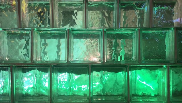 Luminous green glass blocks neon texture background. Close-up of square green glass brick wall glowing with vibrant light, creating wavy water-like reflections.