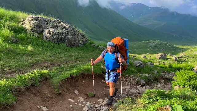A hiker climbs a steep trail through Roshka Valley in a wide alpine view, carrying expedition gear across green slopes beneath cloud-covered peaks, endurance travel