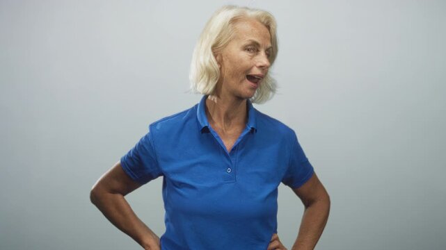 Blonde middleaged woman in blue polo sticking tongue out with hands on hips in studio; playful defiance.