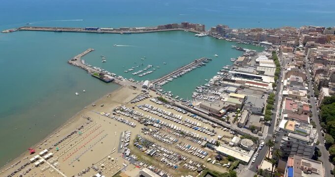 Aerial wide shot of the Anzio harbor and coastline, featuring vibrant sea waters, docked boats, sandy beaches, breakwaters, and urban coastal buildings in a summer day. It's a coastal town in Italy.