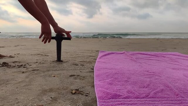 Close up of a brown-skinned woman's hands securing a large beach umbrella anchor into the golden sand, representing summer beach activities, outdoor leisure, vacation preparation