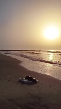 Vertical video of flip flops resting on golden beach sand at the shoreline as gentle waves move in and out, with sea foam repeatedly touching the footwear, creating a peaceful scene