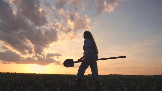 A teenage farmer walks through a field, holding a shovel. The teenage farmer takes in the sunset as clouds drift across the sky.