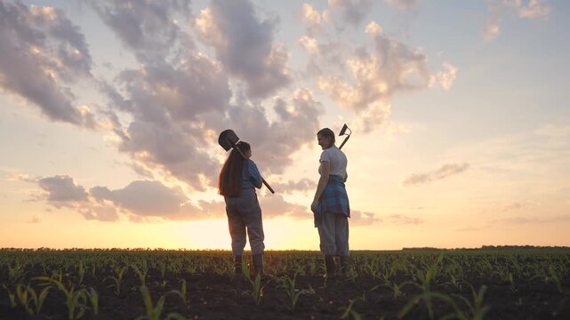 In the field, a woman and a girl stand together, holding tools. The tools reflect their work as they converse against the sunset sky.
