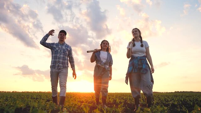 Three individuals walk through a field, carrying tools as they approach the sunset. The man, girl, and woman display a sense of teamwork and community.