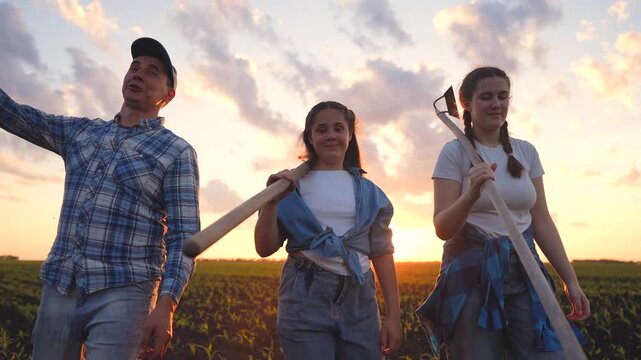 A man, a girl, and a woman walk in a field, carrying tools. The girl smiles as they chat, enjoying their time together in the scenic setting.