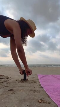 woman on a sandy beach securing a large umbrella anchor into the sand, with the sea visible in the background, representing summer vacation preparation, beach lifestyle, sun protection