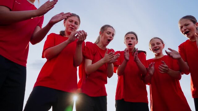 Youth girls soccer team in red jerseys uniting in a pre-match huddle, clapping and encouraging each other during outdoor training under a clear evening sky