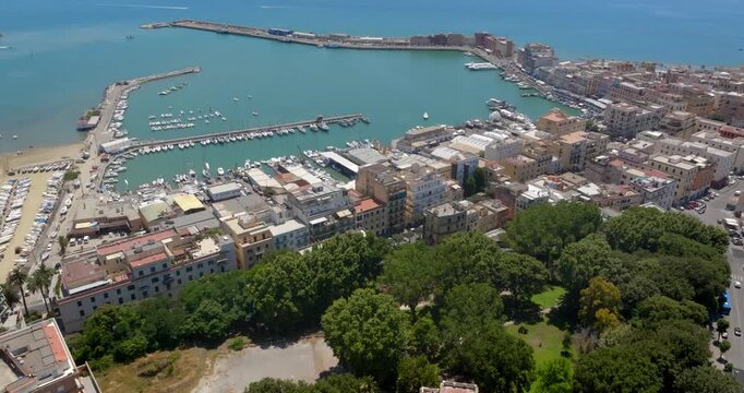 Aerial view of the harbor and coastal town of Anzio, located near Rome in Italy, featuring clear turquoise waters, breakwaters, docks with boats, and densely clustered buildings in a sunny summer day.