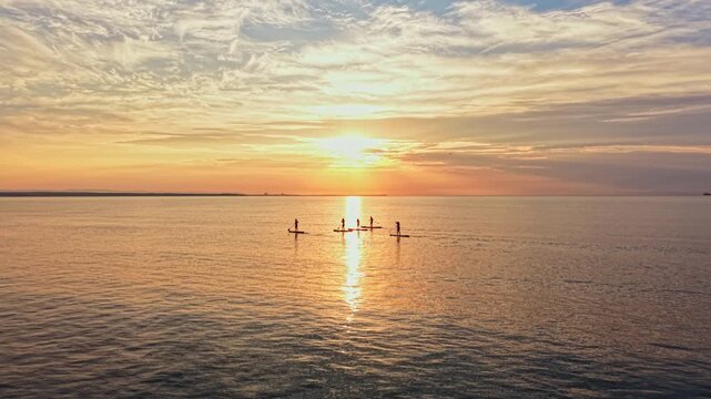 Paddleboarders are on the water during sunset. The sun is low, casting reflection on the surface. Five people can be seen paddling in a line as colors fill the sky.