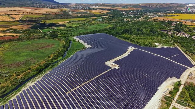 Solar panels are arranged in rows on a hillside close to a river. Fields and hills surround the area under clear blue skies. The scene shows renewable energy efforts in action.