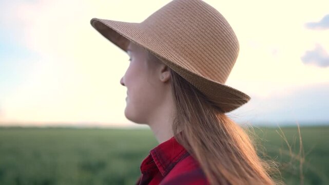 The young woman stands in the field, wearing a large hat. The woman gazes toward the horizon, surrounded by a green field and soft light.
