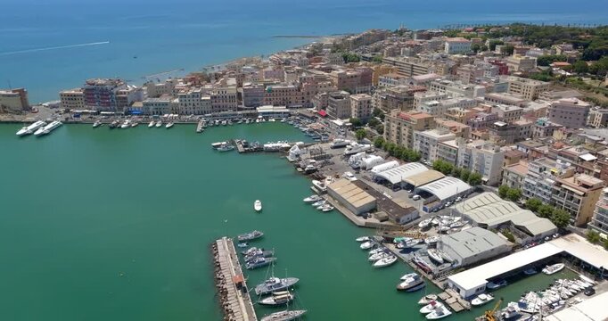 Aerial view of the coastal town of Anzio, near Rome in Italy, showing the bustling port with boats and yachts, turquoise sea waters, breakwaters, and colorful urban buildings along the coastline.